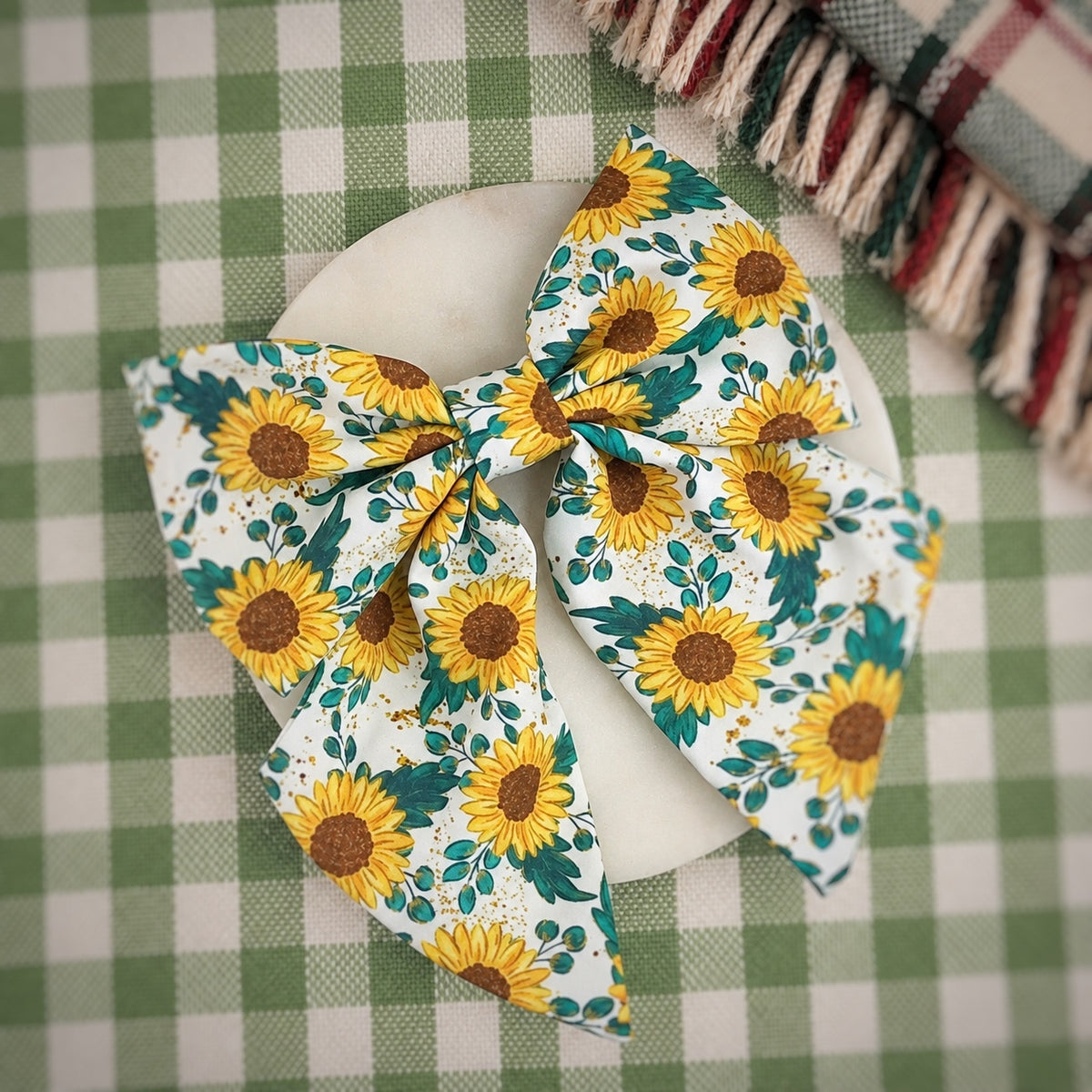 A large fabric hair bow with a yellow sunflower and green leaf pattern, displayed on a white oval plate over a green and white checkered tablecloth.