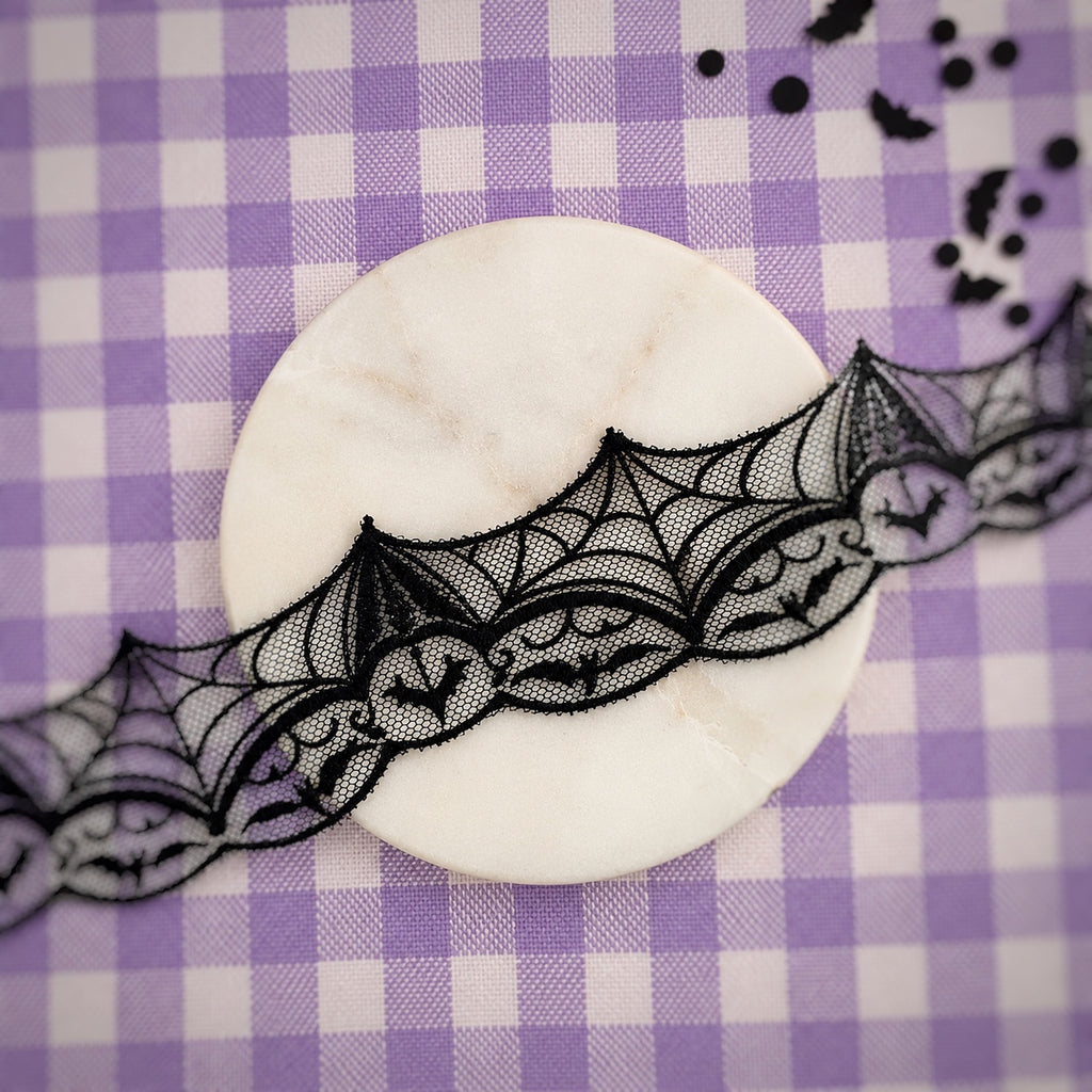 Black lace ribbon with a bat motif draped over a round white marble coaster on a purple and white checkered background.