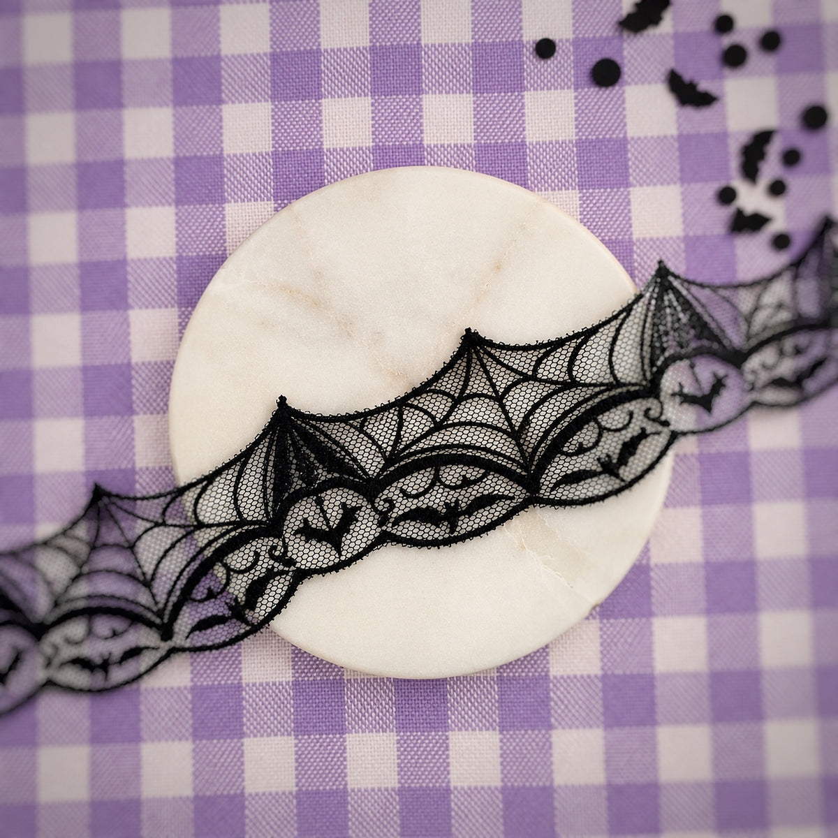 Black lace ribbon with a bat motif draped over a round white marble coaster on a purple and white checkered background.