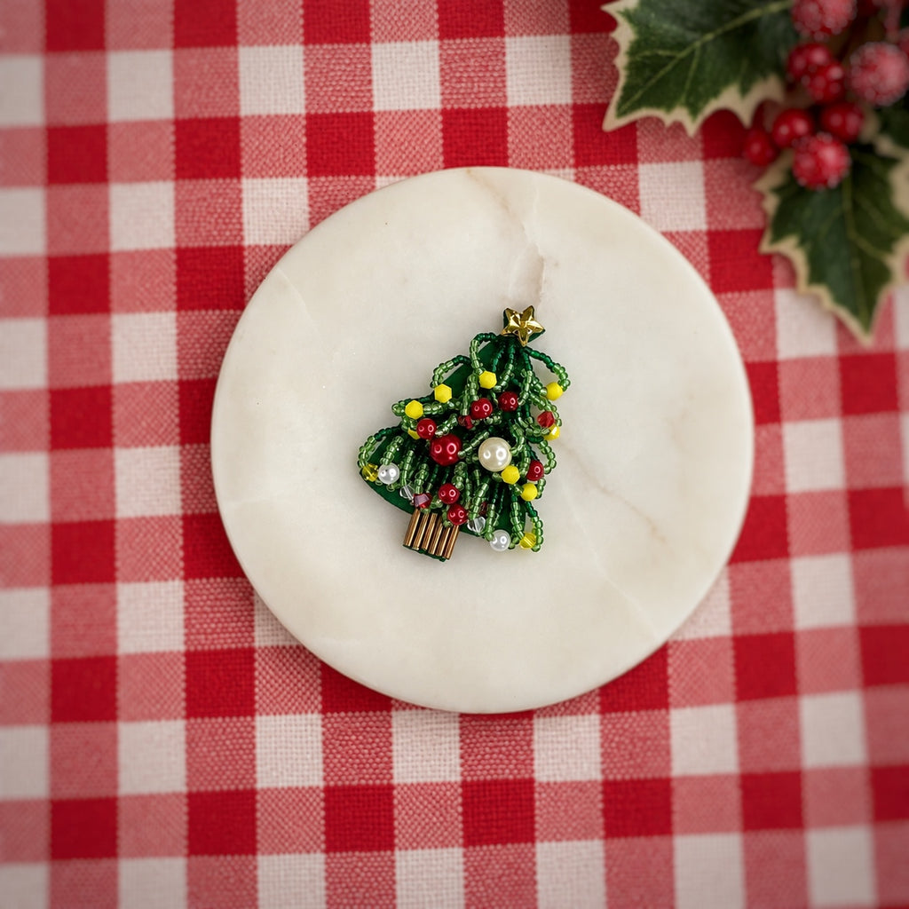 A small Christmas tree enamel pin with green leaves, red and yellow berries, and white pearl-like ornaments, displayed on a round white marble coaster over a red and white checkered tablecloth with holly in the corner.