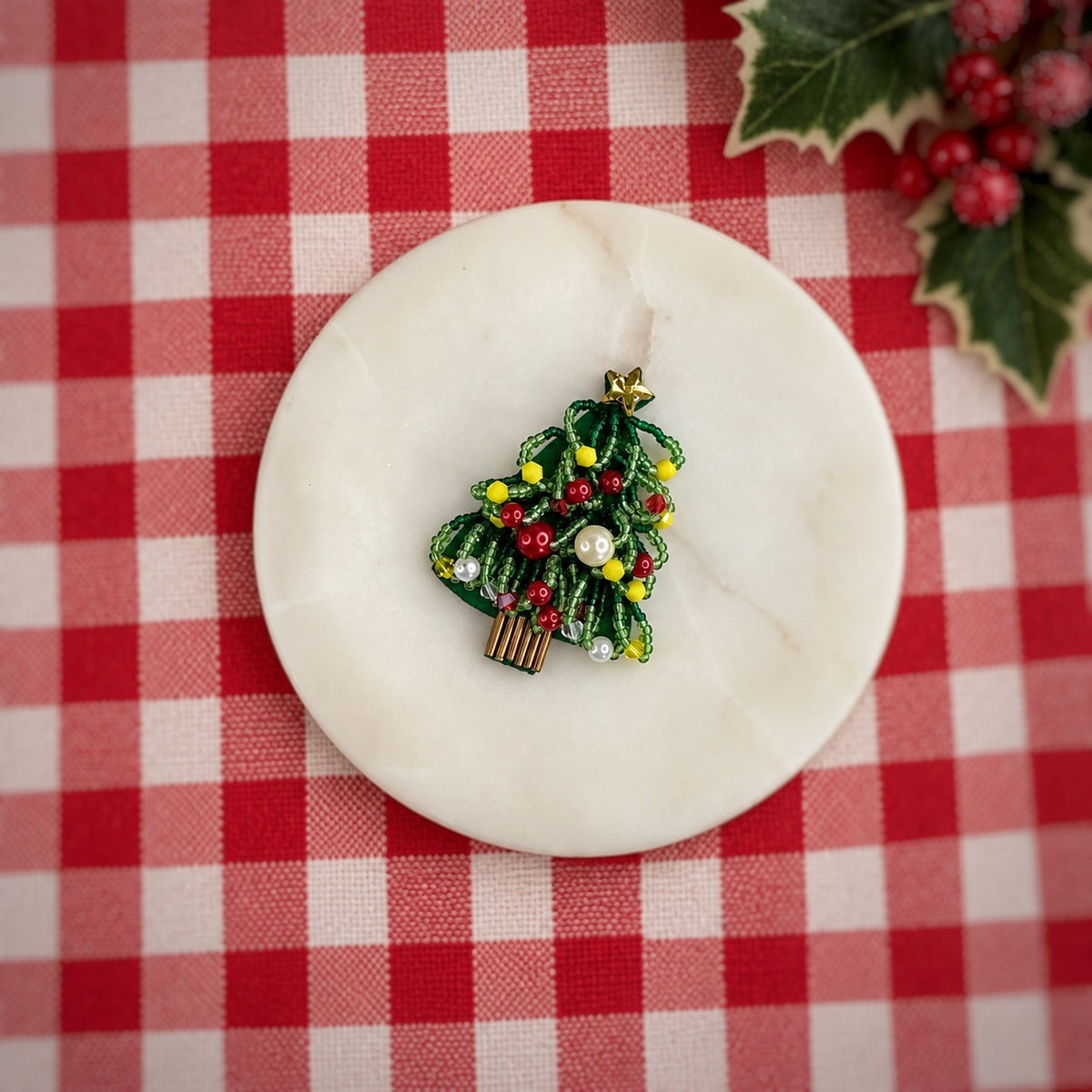 A small Christmas tree enamel pin with green leaves, red and yellow berries, and white pearl-like ornaments, displayed on a round white marble coaster over a red and white checkered tablecloth with holly in the corner.