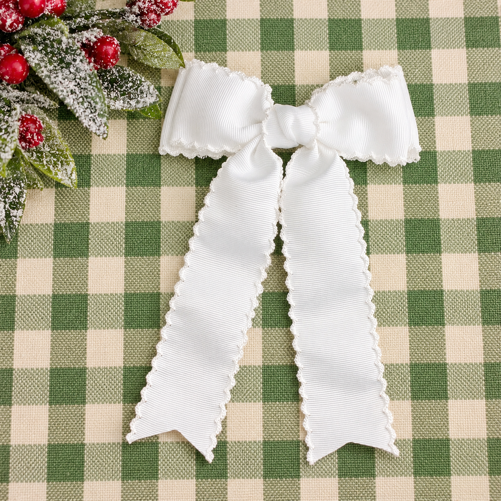 A white grosgrain ribbon bow with scalloped edges, laid flat on a green and beige checkered fabric background with a decorative holly sprig in the top-left corner.