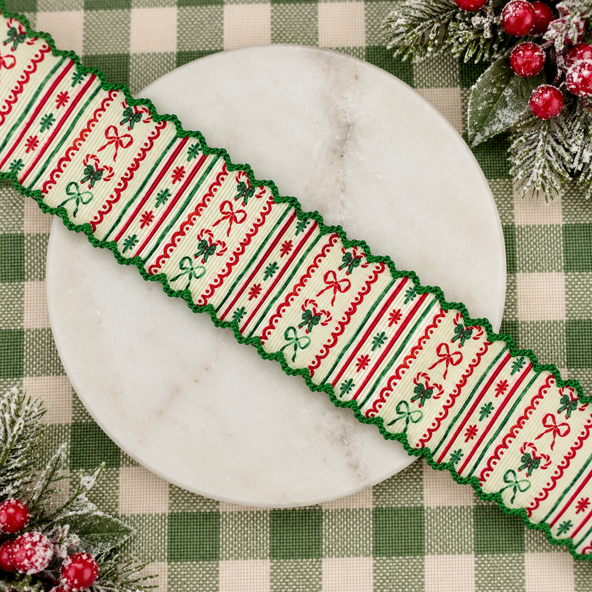 A festive Christmas-themed decorative ribbon with a moonstitch scalloped edge, laid diagonally across a round white marble plate. The ribbon features a cream background with red and green stripes, bows, holly leaves, and snowflake patterns.