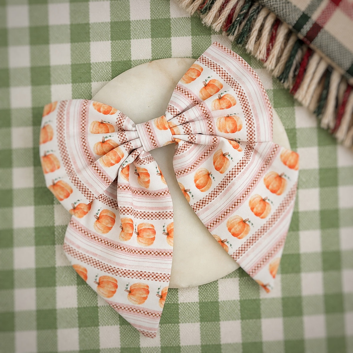 A large bow hair clip featuring a pumpkin and stripe pattern in orange, brown, and white, displayed on a round white plate over a green and white checkered tablecloth.