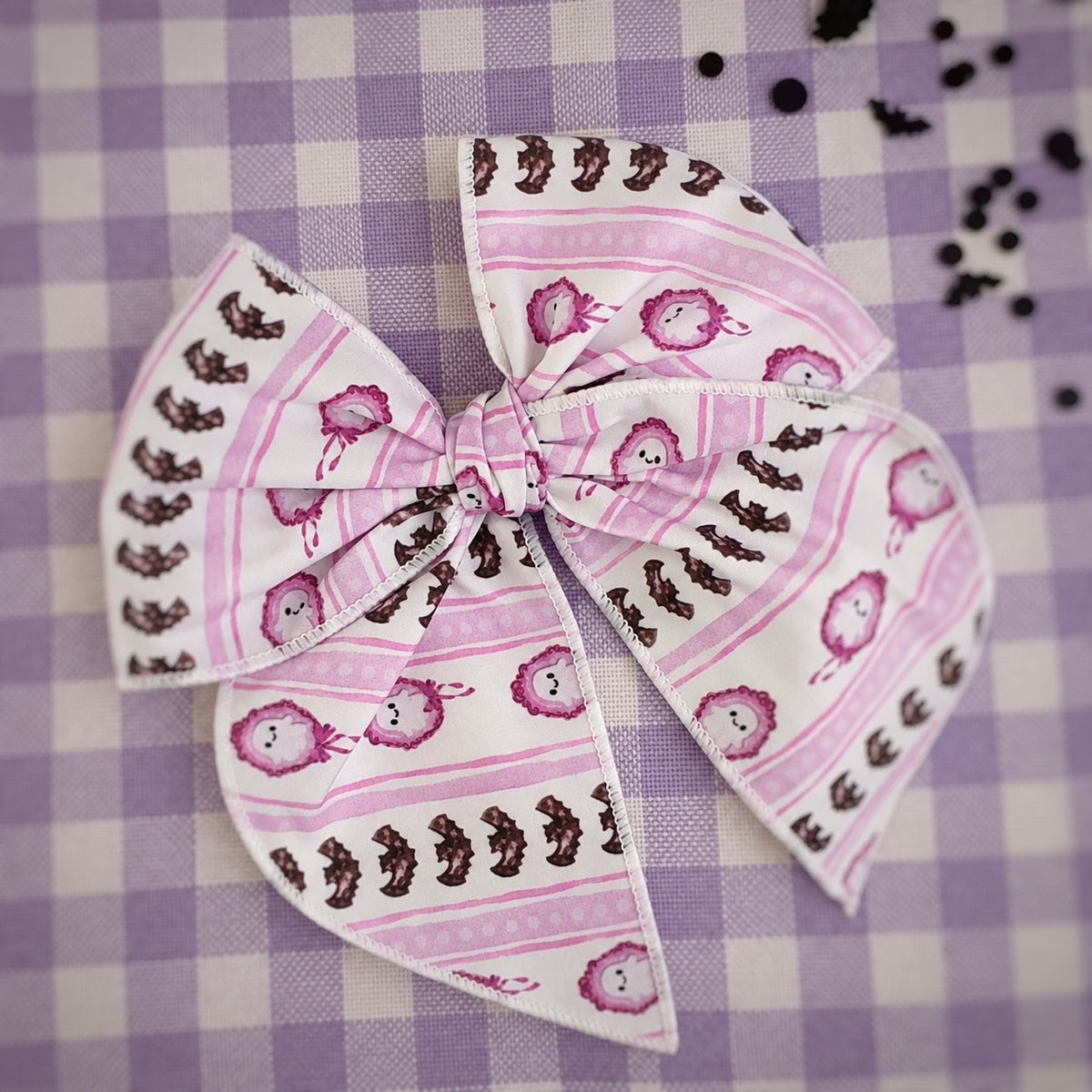 A fabric hair bow featuring a pattern of ghosts, pink stripes, and bat prints on a white background, styled on a purple and white checkered fabric surface.