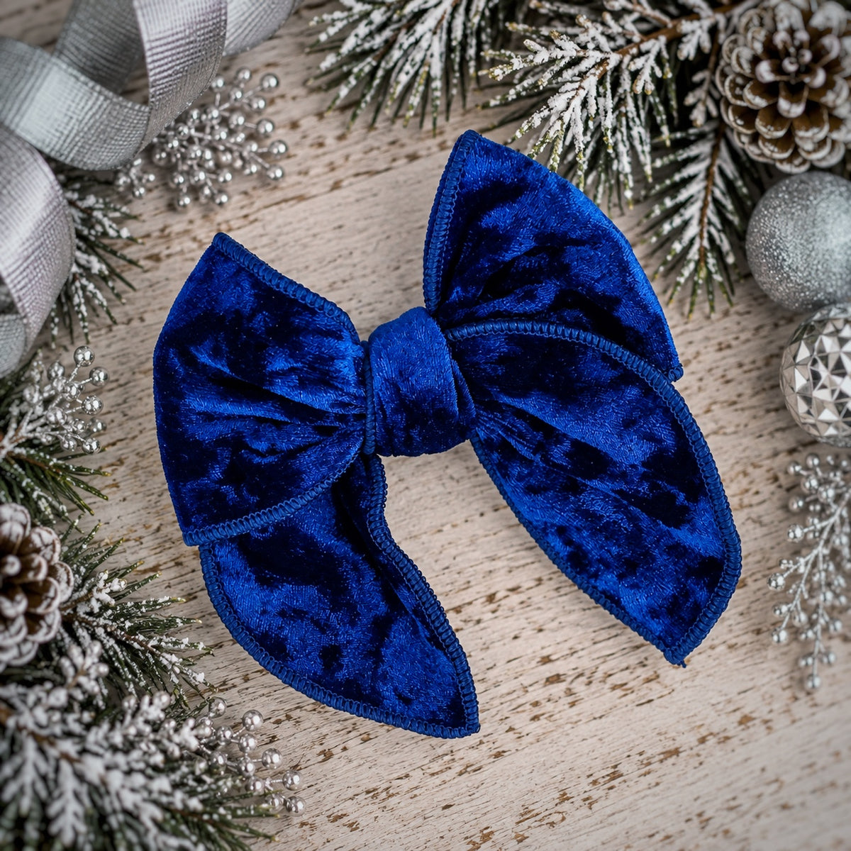 A royal blue velvet hair bow with stitched edges, styled on a light wood surface with festive holiday decorations including pine branches, silver ornaments, and a ribbon.