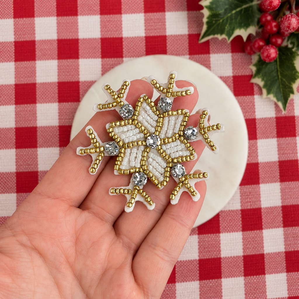 A hand holds a decorative snowflake bow center made of white and gold seed beads with clear rhinestones, set against a red and white checkered tablecloth with holly leaves in the corner.