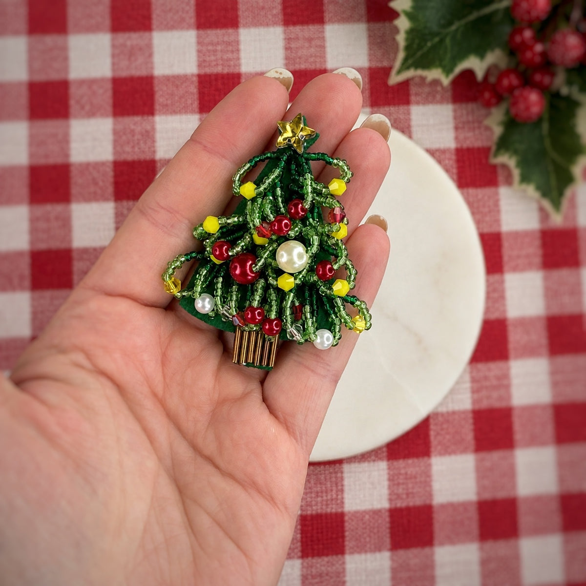 A hand holds a small Christmas tree brooch with green glittery branches, red and yellow beads, white pearl-like ornaments, and a gold star on a red-and-white checkered tablecloth background.