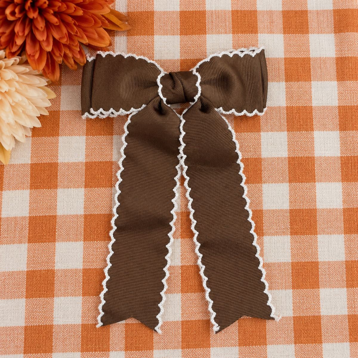 A brown grosgrain ribbon bow with white scalloped edges, laid flat on an orange and white checkered fabric background with decorative flowers in the top left corner.