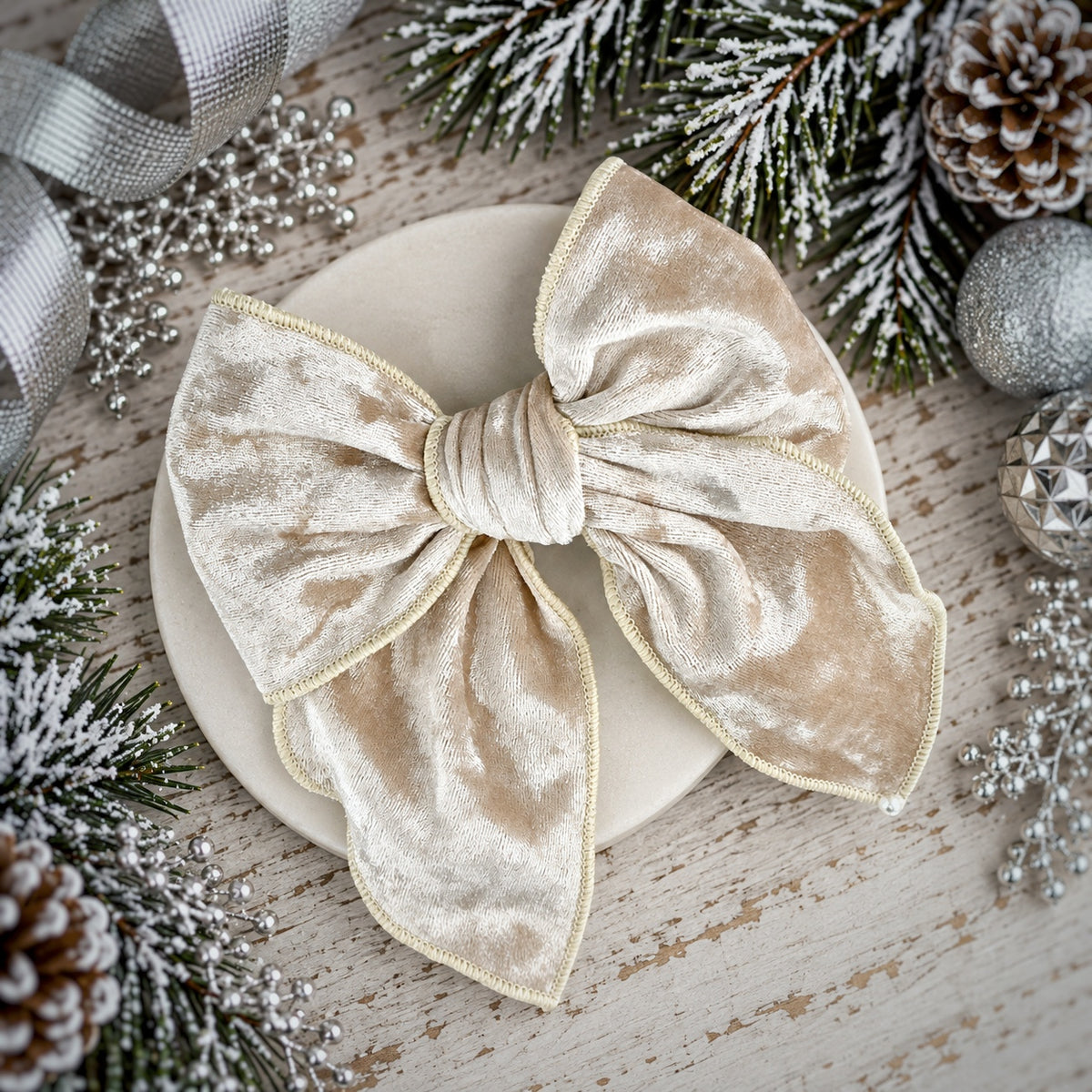 A large champagne beige velvet bow with a gold satin trim, displayed on a round cream-colored plate. The plate is set against a rustic wooden background decorated with pine branches, pinecones, and silver holiday ornaments.