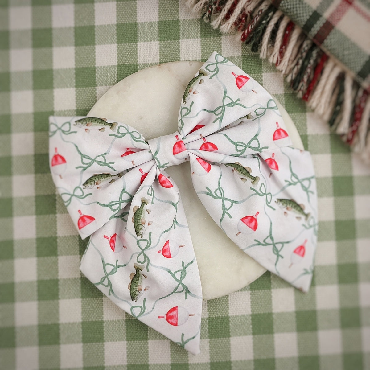 A fabric hair bow with a white background and a print of green bows, red bobbers, and green fish, displayed on a round white dish over a green and white checkered tablecloth.