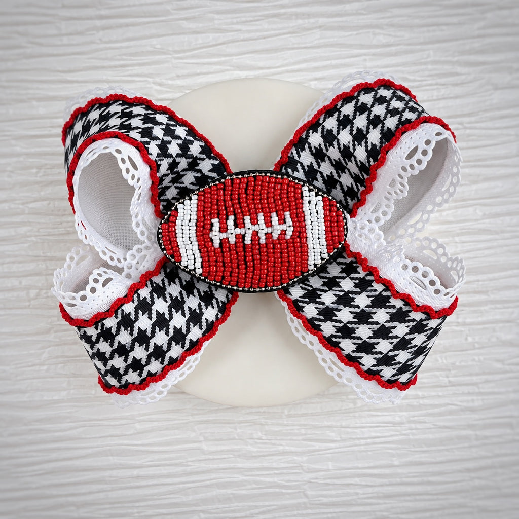 A decorative hair bow featuring a black and white houndstooth pattern, red and white lace trim, and a central beaded football motif in red, white, and black, displayed on a white dish against a light wood background.