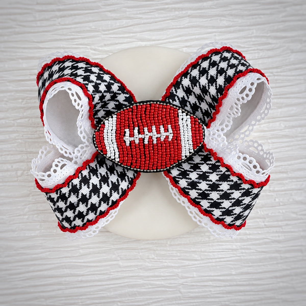 A decorative hair bow featuring a black and white houndstooth pattern, red and white lace trim, and a central beaded football motif in red, white, and black, displayed on a white dish against a light wood background.