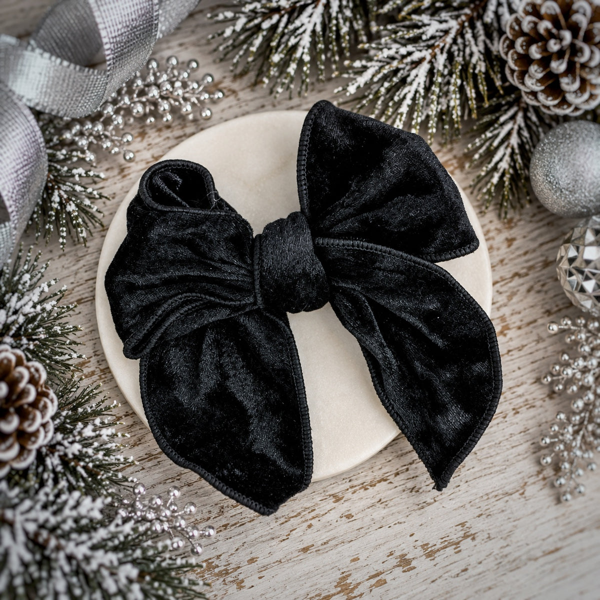 A large black velvet bow displayed on a round cream-colored coaster, surrounded by festive holiday decorations including pinecones, silver ribbon, and snow-dusted evergreen branches on a rustic wooden surface.