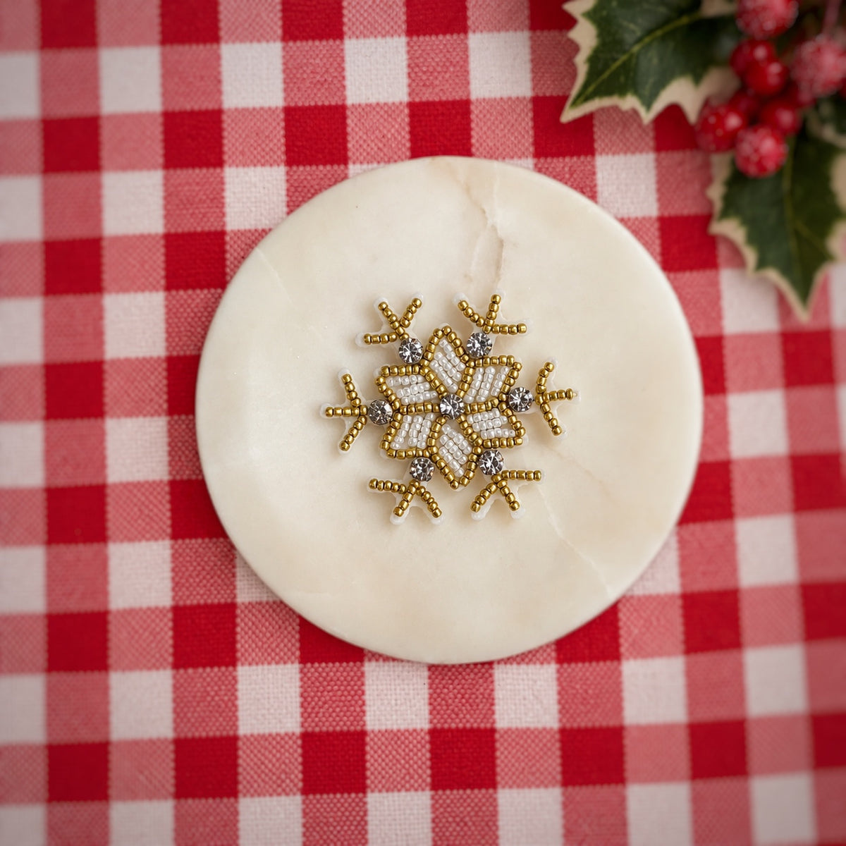 A gold and white beaded snowflake bow center displayed on a round white marble coaster, set against a red and white checkered holiday tablecloth with holly leaves in the corner.