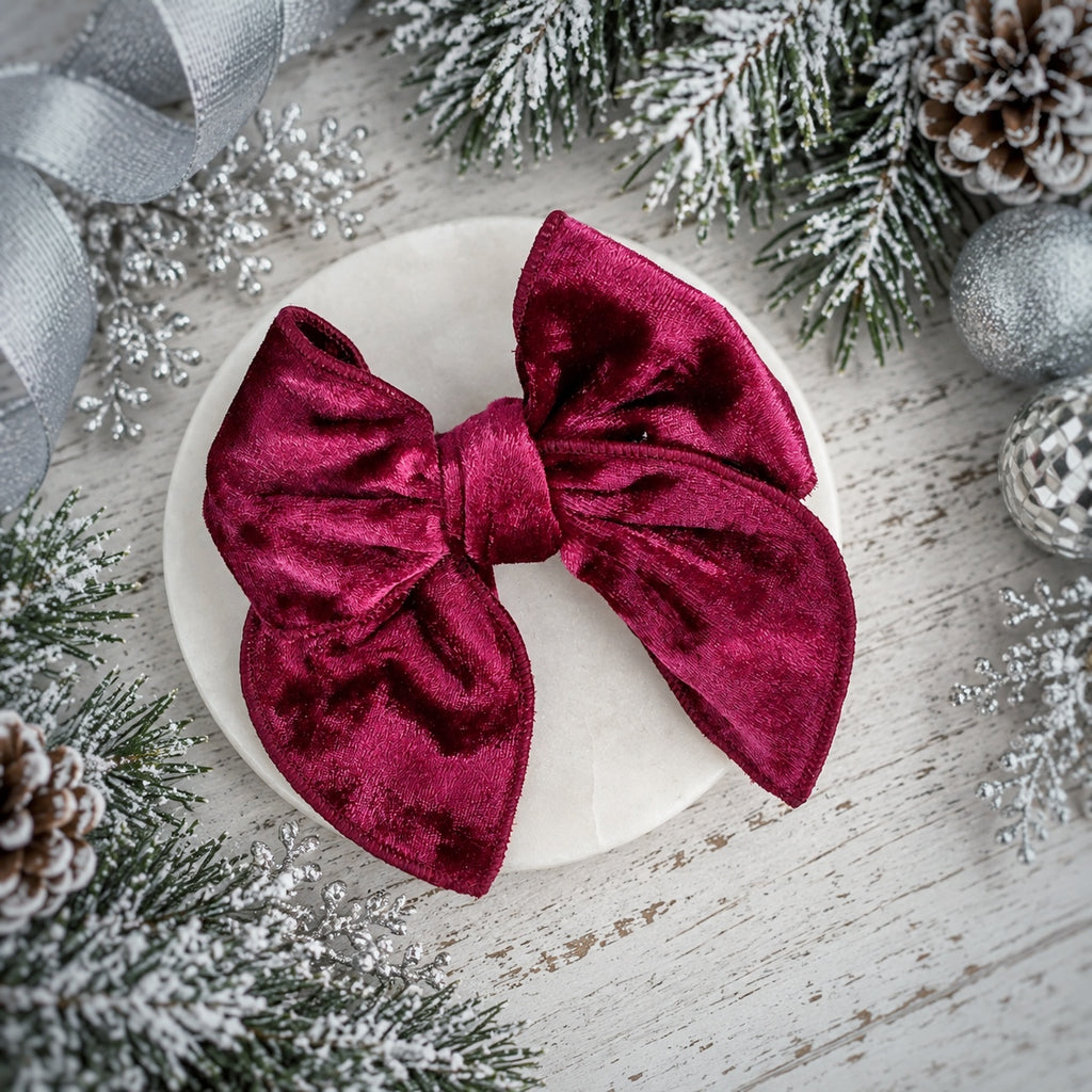 A large deep burgundy velvet bow displayed on a round white plate, surrounded by festive Christmas decorations including pine branches, silver ornaments, and a silver ribbon on a whitewashed wooden surface.