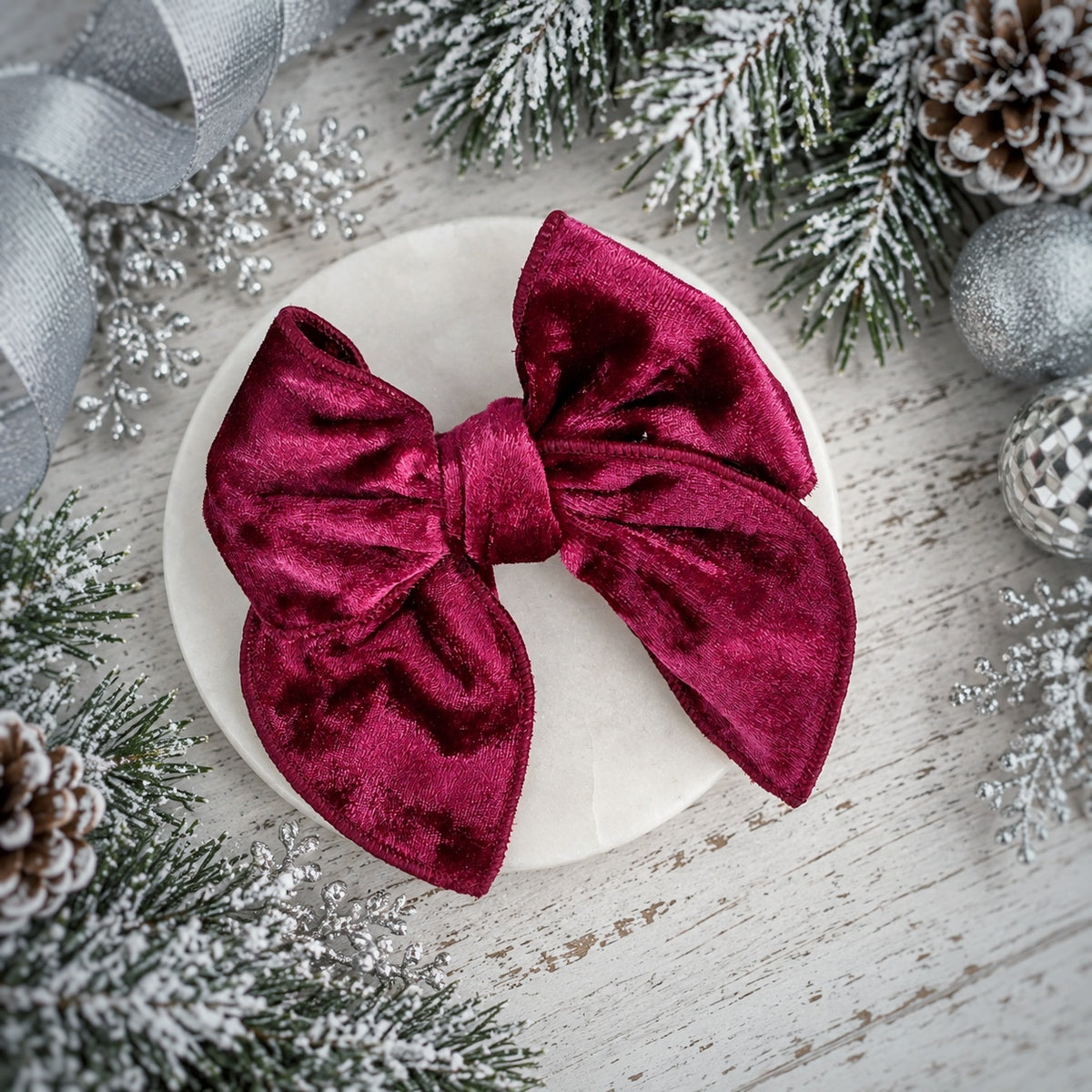 A large deep burgundy velvet bow displayed on a round white plate, surrounded by festive Christmas decorations including pine branches, silver ornaments, and a silver ribbon on a whitewashed wooden surface.