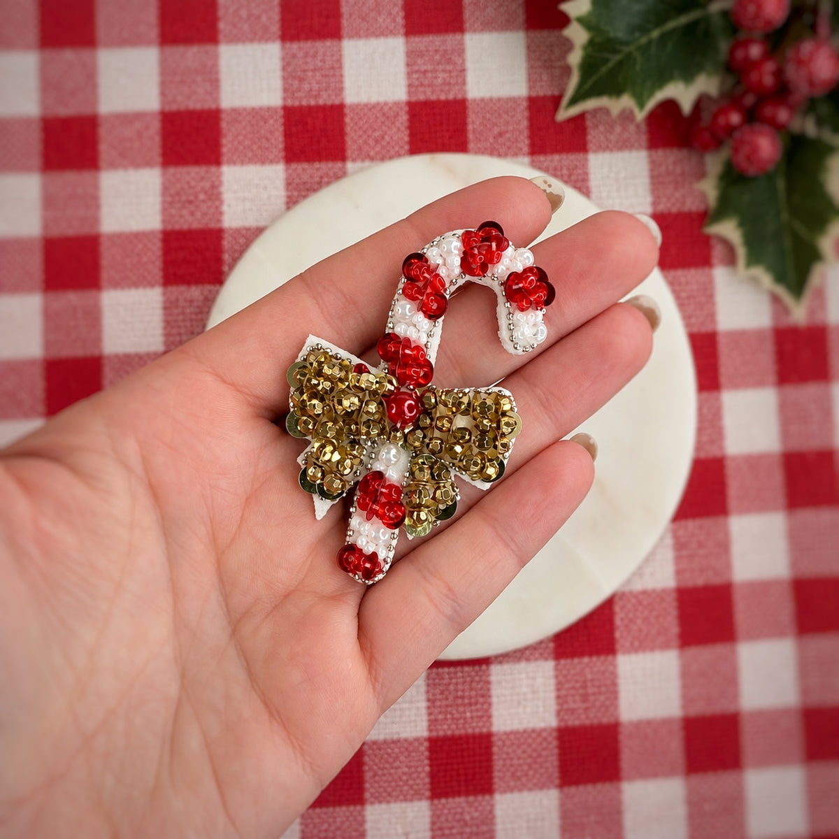 A hand holds a decorative Christmas-themed brooch featuring a white sugar-cane motif with red berry accents and a gold bow embellished with red beads, set against a red and white checkered tablecloth with holly leaves in the corner.