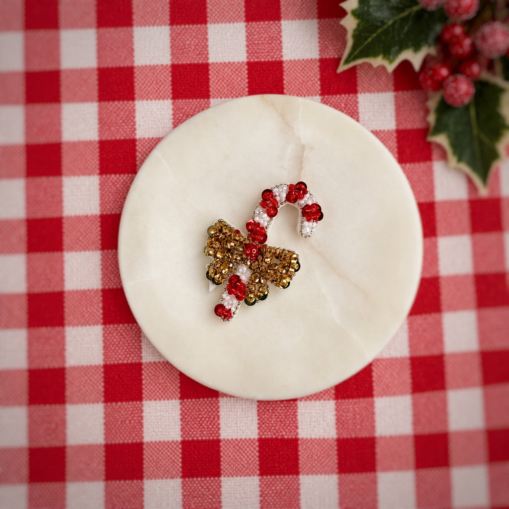 A holiday-themed beaded ornament or decorative piece shaped like a candy cane and bow, featuring red, white, and gold beads, displayed on a round white marble coaster over a red and white checkered tablecloth with holly leaves in the corner.