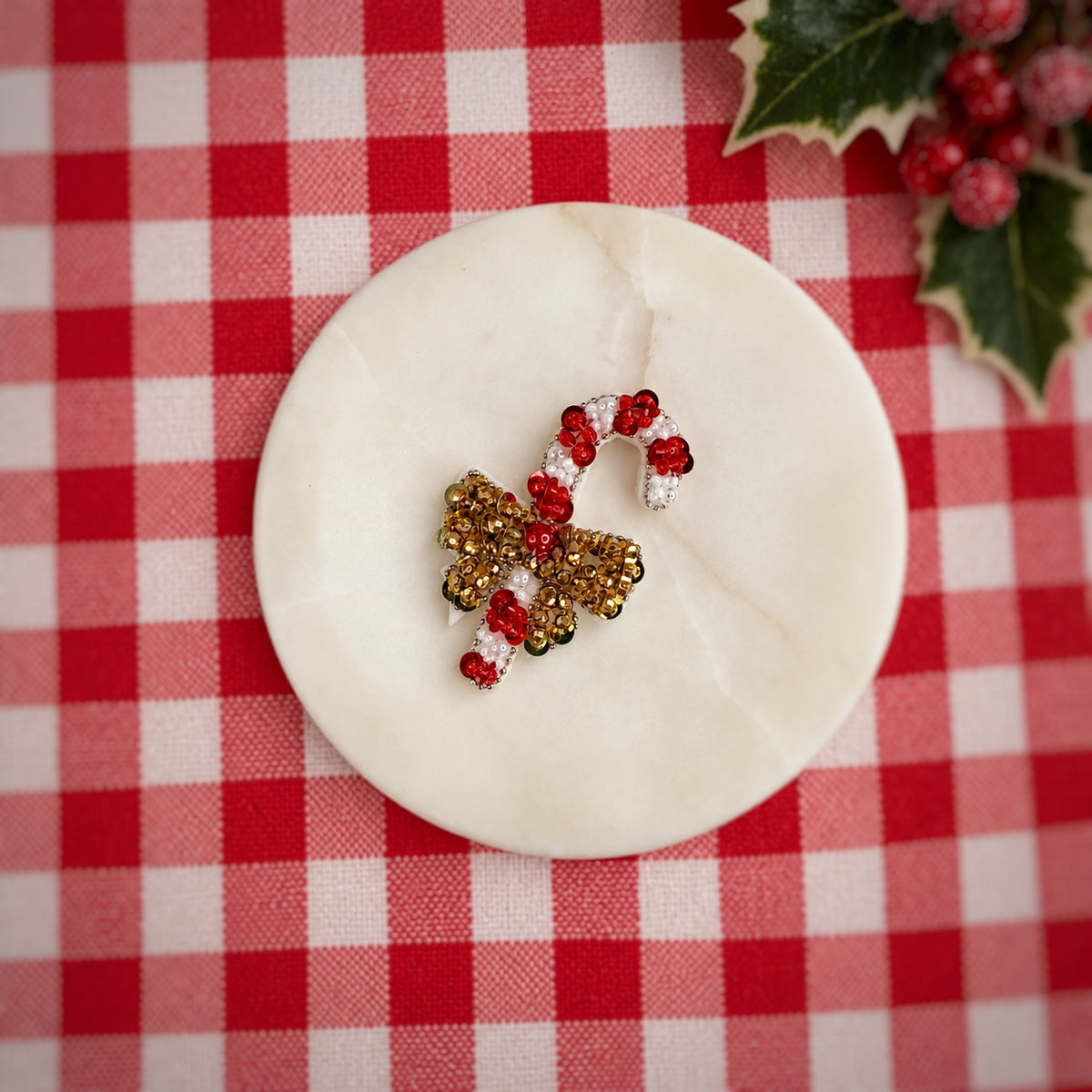 A holiday-themed beaded ornament or decorative piece shaped like a candy cane and bow, featuring red, white, and gold beads, displayed on a round white marble coaster over a red and white checkered tablecloth with holly leaves in the corner.