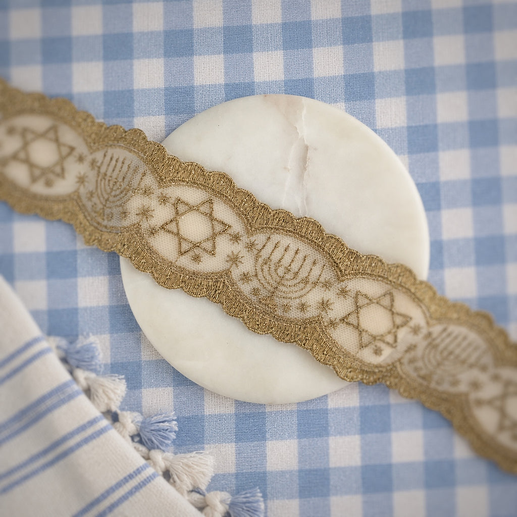 A close-up of a gold embroidered decorative trim with Star of David and menorah motifs, laid diagonally across a round white stone coaster on a blue and white checkered fabric surface.