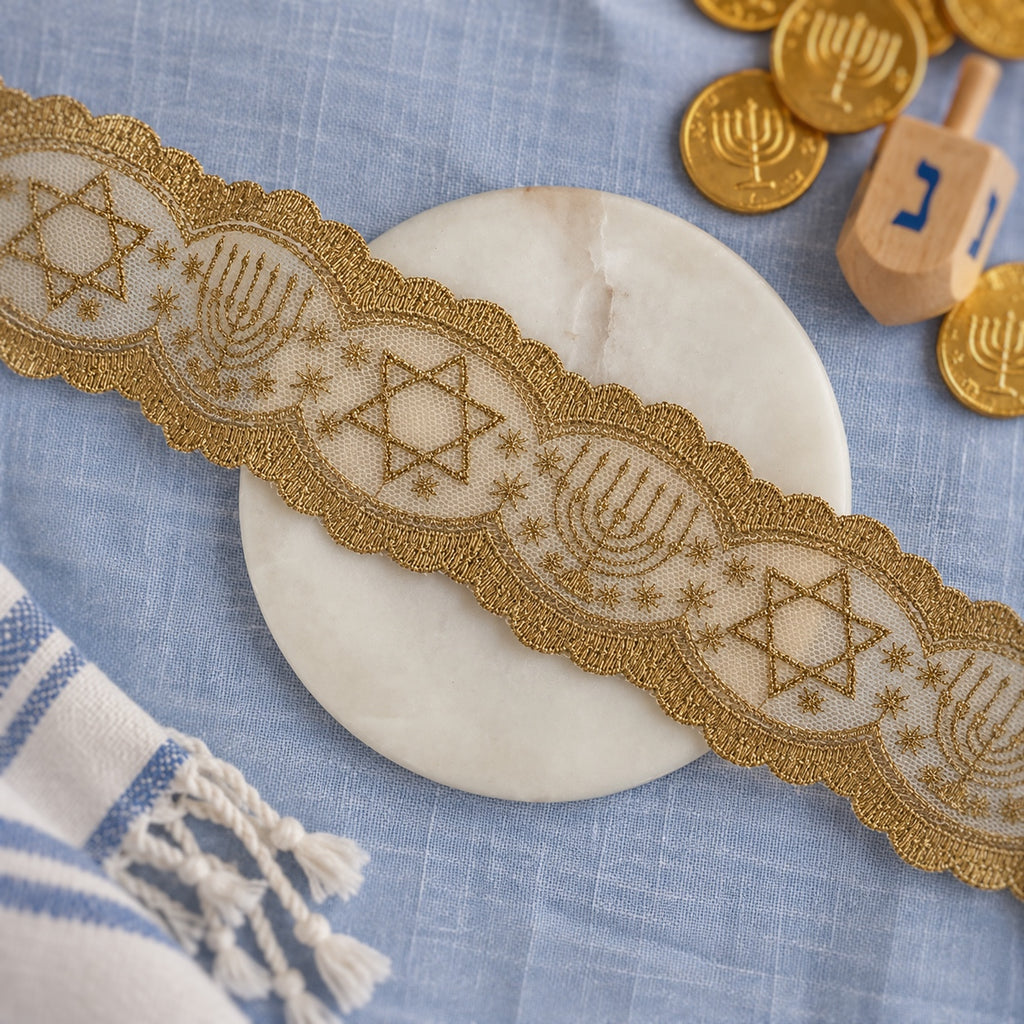 A close-up of a decorative embroidered lace trim with gold-tone stitching and Star of David and Menorah motifs, laid diagonally across a round white stone coaster on a light blue fabric surface. A striped textile, gold coins, and a wooden dreidel are partially visible as styled props.