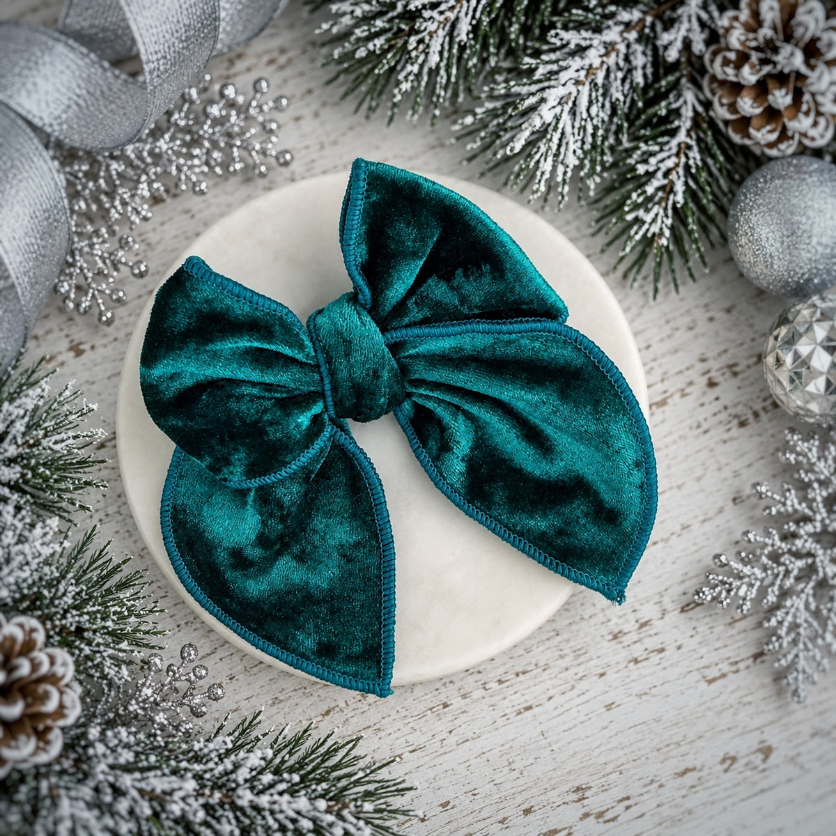 A large teal velvet bow with stitched edges, displayed on a round white dish surrounded by festive holiday decorations including pine branches, silver ornaments, and a ribbon.
