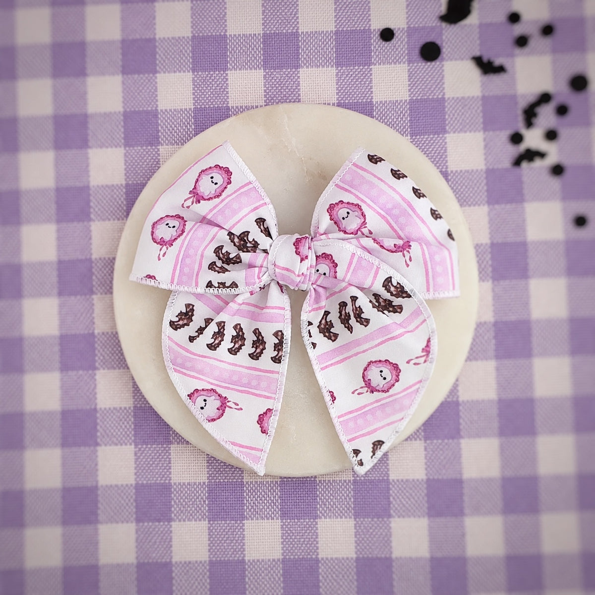 A fabric hair bow with a pink and white striped pattern, featuring ghosts and bat silhouettes, displayed on a round wooden coaster over a purple and white checkered tablecloth.