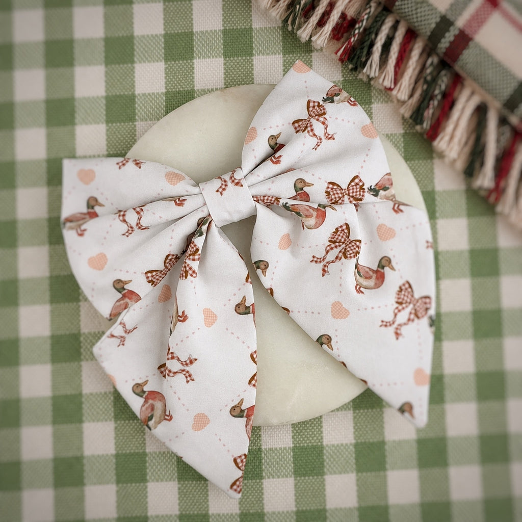 A large fabric hair bow with a white background and a pattern of brown birds, checkered bows, and small peach hearts, displayed on a round white dish over a green and white checkered tablecloth.