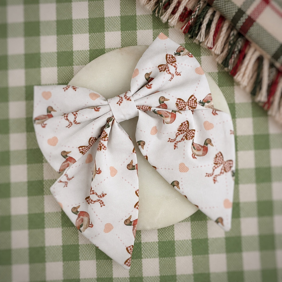 A large fabric hair bow with a white background and a pattern of brown birds, checkered bows, and small peach hearts, displayed on a round white dish over a green and white checkered tablecloth.
