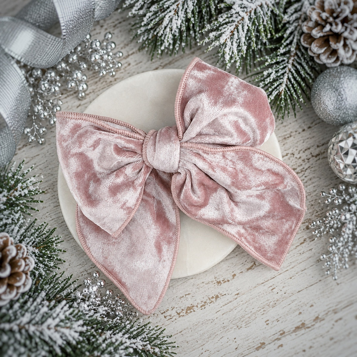 A dusty pink velvet bow with a subtle marbled texture, displayed on a round white plate surrounded by festive holiday decorations including pine branches, silver ornaments, and a ribbon.