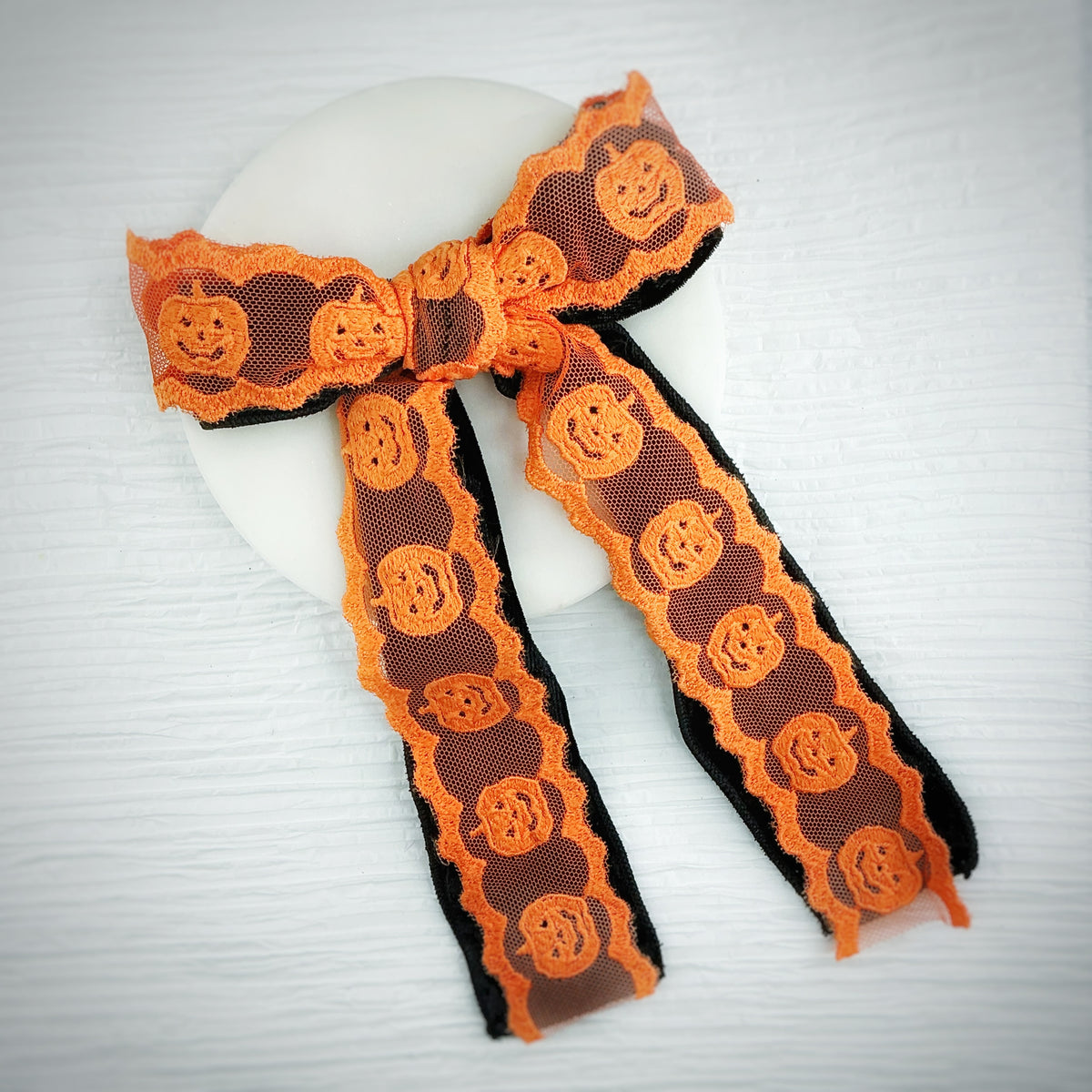 An orange and black lace ribbon bow with a pumpkin pattern, displayed on a white circular stand against a light gray wood-textured background.