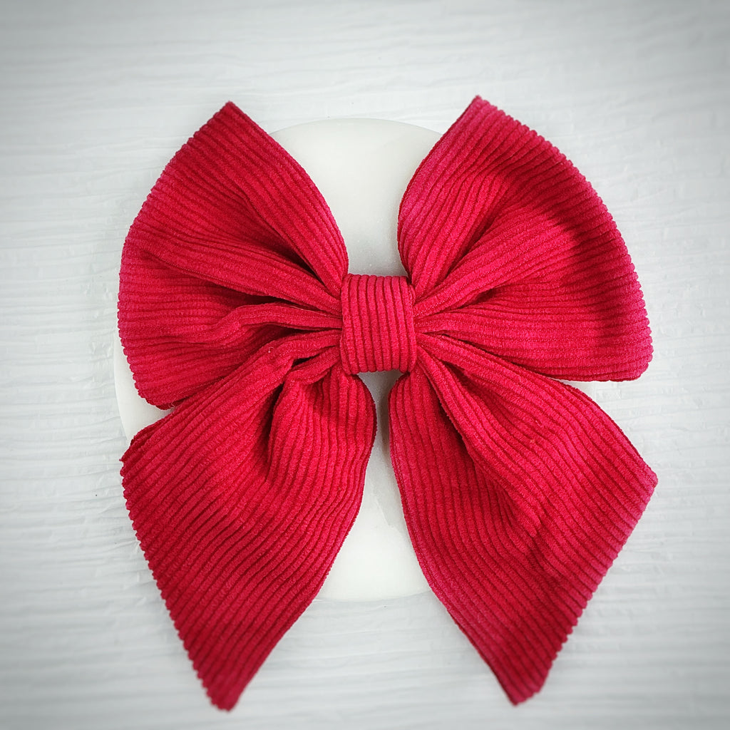 A large red corduroy bow displayed on a white circular stand against a light gray textured background.
