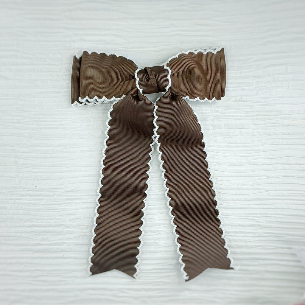 A brown grosgrain ribbon bow with white scalloped edges, tied neatly on a textured white background.