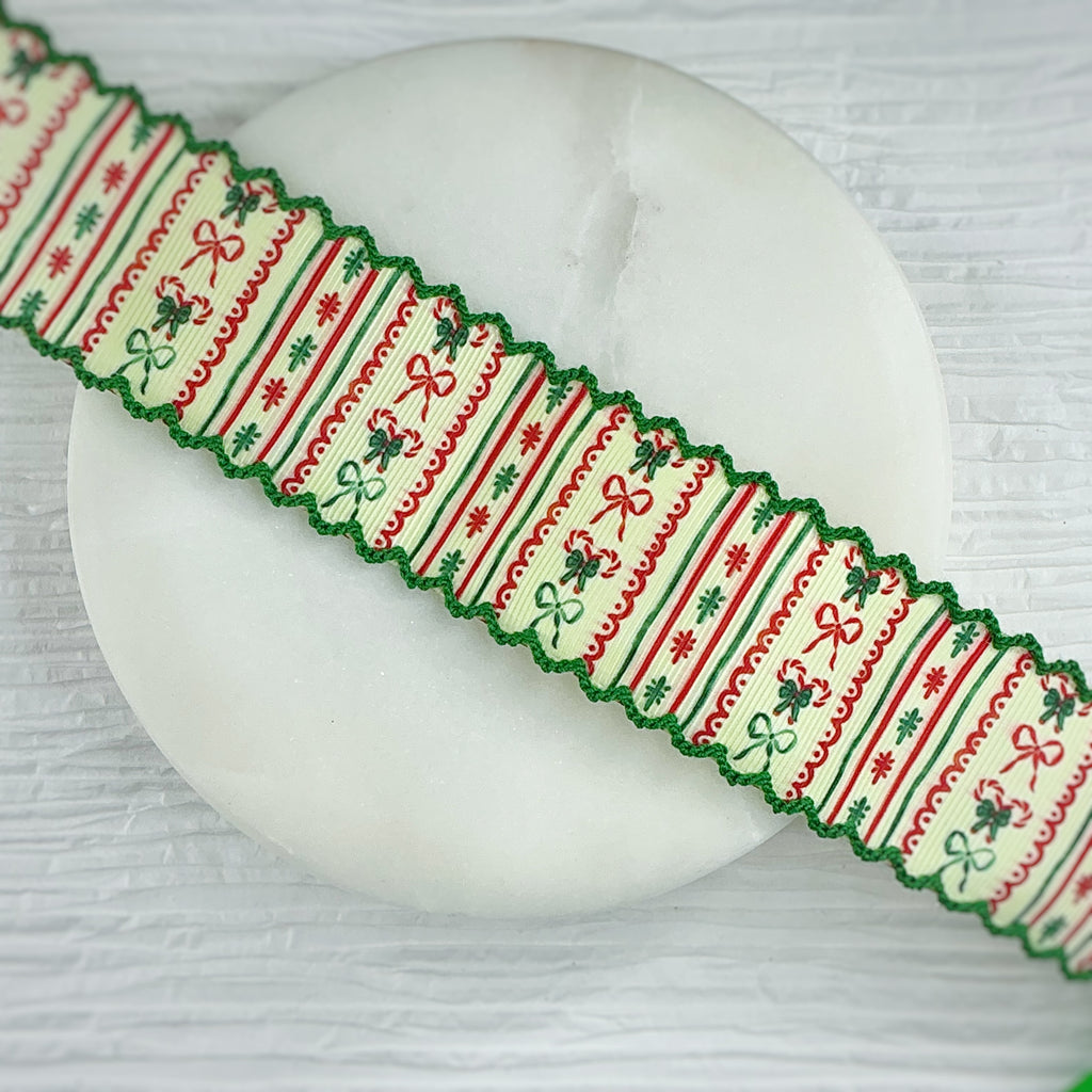A festive Christmas-themed decorative ribbon with a moonstitch scalloped edge, laid diagonally across a round white marble plate. The ribbon features a cream background with red and green stripes, bows, holly leaves, and snowflake patterns.