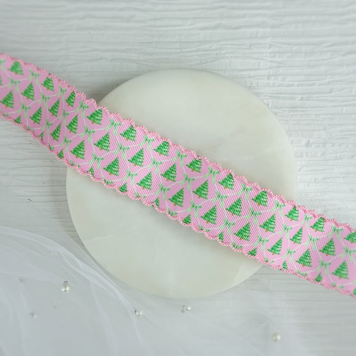 A pink grosgrain ribbon with a repeating green Christmas tree pattern and a scalloped edge, draped diagonally across a round white display stand on a white wood-textured surface.