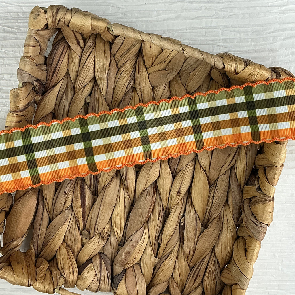 A close-up of a woven natural fiber basket with a decorative ribbon trim across the top. The ribbon has a plaid pattern in orange, olive green, brown, and white, with orange fringe on the sides.