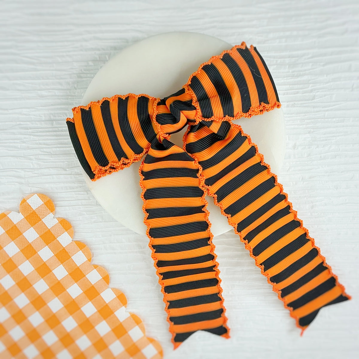 An orange and black striped grosgrain ribbon bow with a moonstitch edge, styled on a white circular plate with a matching orange and white gingham ribbon prop on a white textured background.