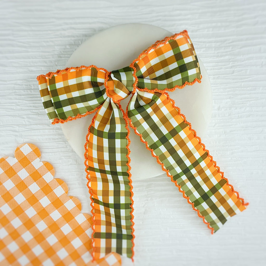 An orange and green plaid ribbon bow with scalloped edges, displayed on a white oval plate over a white wood background, with a matching plaid ribbon scrap piece in the lower left corner.