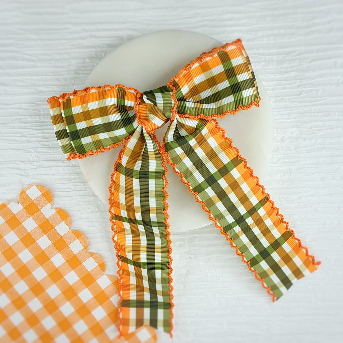 An orange and green plaid ribbon bow with scalloped edges, displayed on a white oval plate over a white wood background, with a matching plaid ribbon scrap piece in the lower left corner.