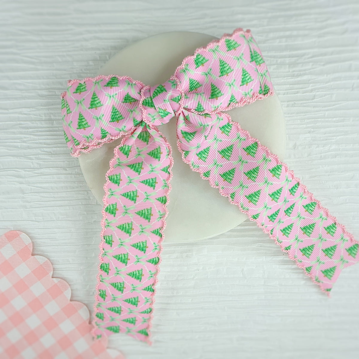 A pink grosgrain ribbon with a green Christmas tree pattern and scalloped edges, tied into a bow on a white circular display stand.