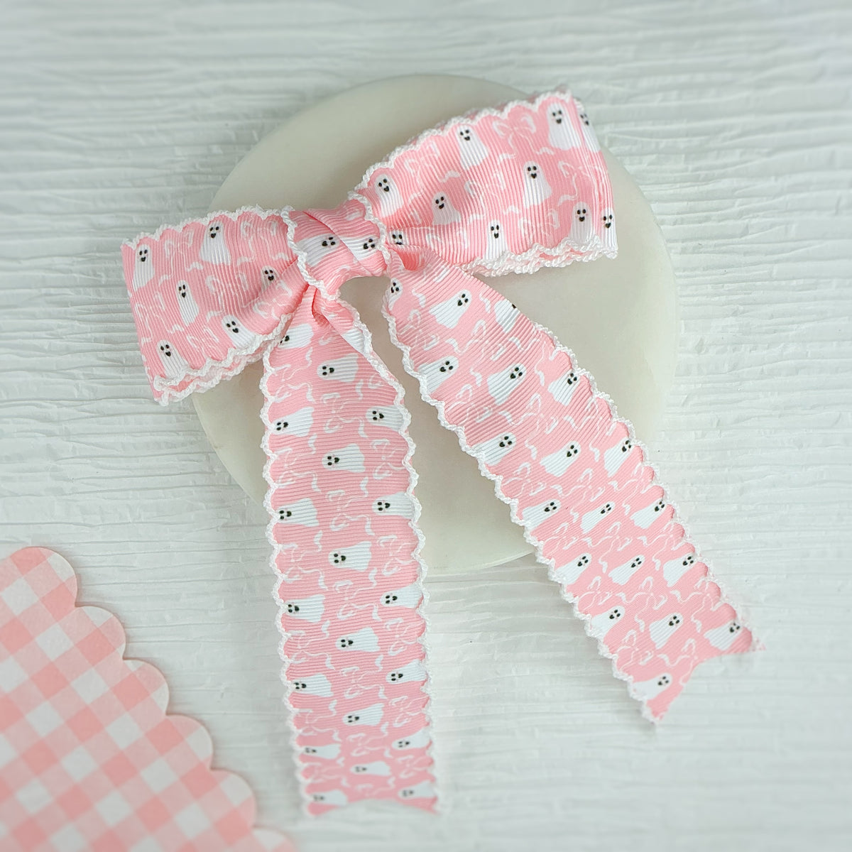 A pink ribbon bow with a repeating ghost print and white scalloped edges, tied in a large bow shape and draped over a round white dish on a light textured surface.