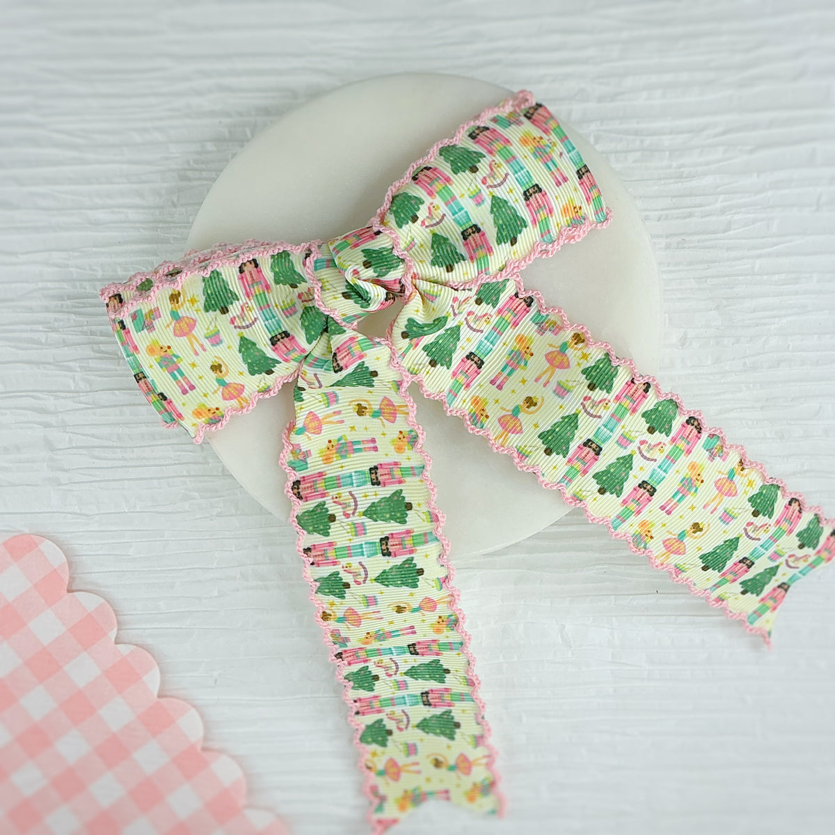 A decorative ribbon bow with a pastel pattern of Christmas trees, presents, and ballerinas, finished with pink scalloped edges, displayed on a white circular stand.