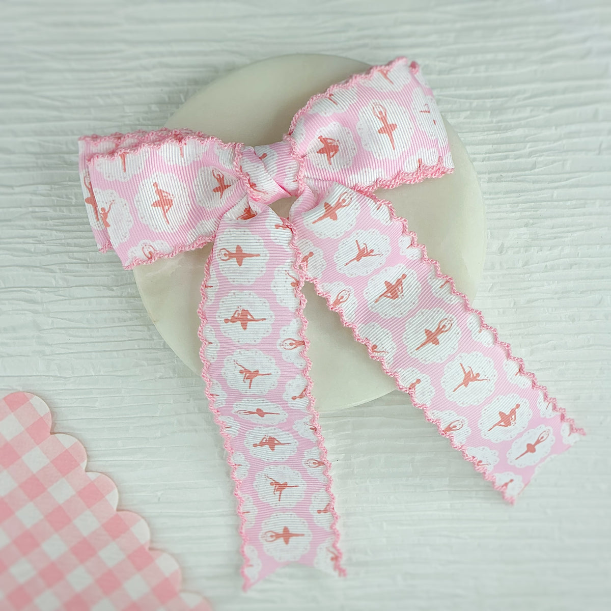 A pink ribbon bow with a scalloped edge, featuring a repeating pattern of ballerina figures inside white hearts, displayed on a round white object against a light textured background.