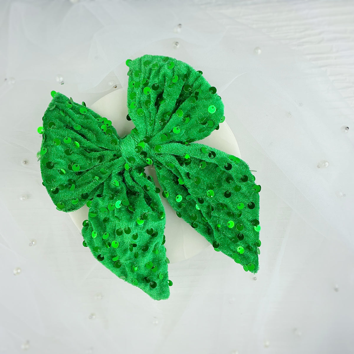 A bright green sequined fabric bow with textured, plush loops, displayed on a white circular stand against a white fabric backdrop with subtle pearl-like embellishments.
