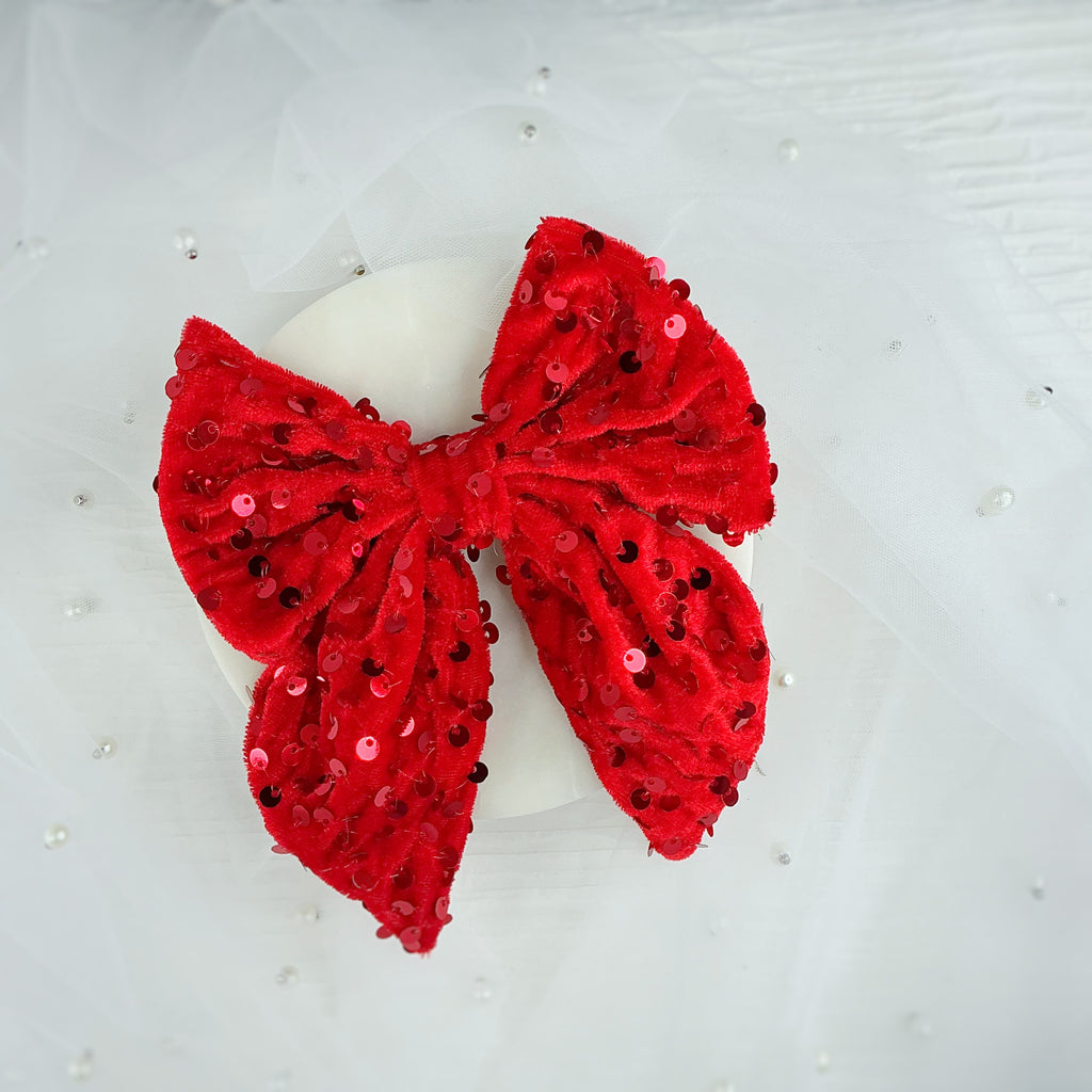 A large red sequined bow with a plush velvet texture, displayed on a white circular stand against a white tulle fabric background.
