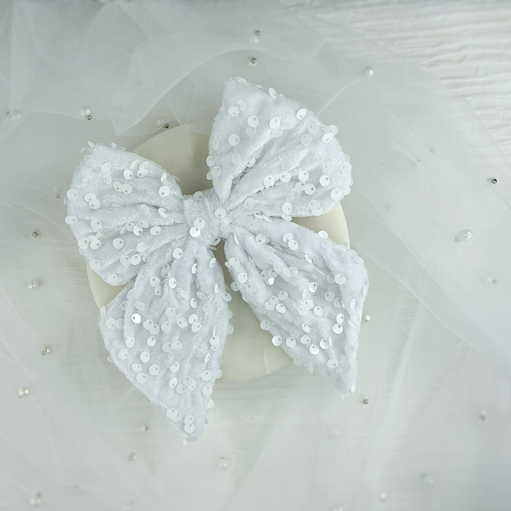A white fabric bow embellished with small silver sequins, displayed on a cream-colored circular stand against a white tulle backdrop with scattered pearl-like beads.