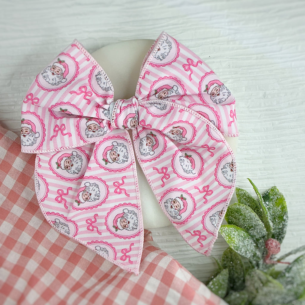 A large fabric hair bow with a pink and white striped pattern featuring cartoon Santa  characters in circular frames, displayed on a white mannequin head with a red-and-white gingham cloth and green foliage props.