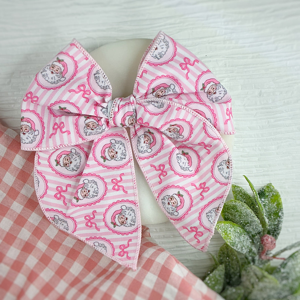 A large fabric hair bow with a pink and white striped pattern featuring cartoon Santa  characters in circular frames, displayed on a white mannequin head with a red-and-white gingham cloth and green foliage props.