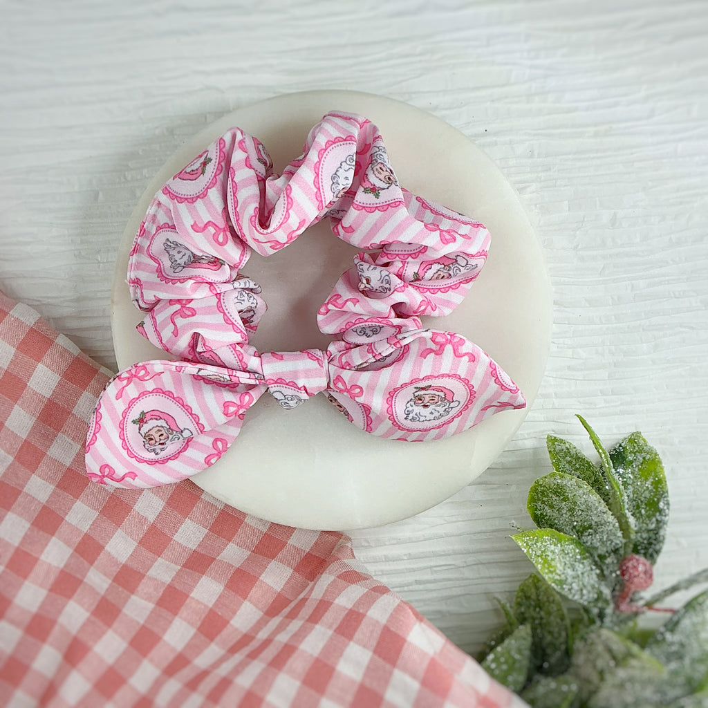Two pink fabric hair scrunchies with a cartoon Santa  pattern, displayed on a round white dish with a pink-and-white checkered cloth and small green leaves in the corner.
